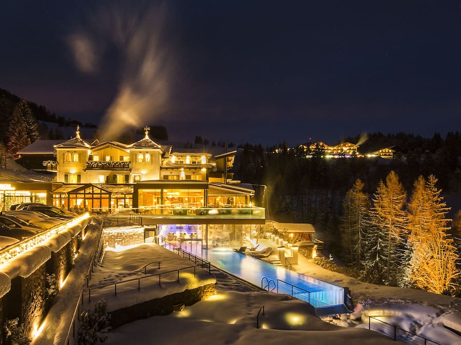 Ein großes, beleuchtetes Hotel mit Schwimmbad, umgeben von schneebedeckten Bäumen und einer Bergkulisse.