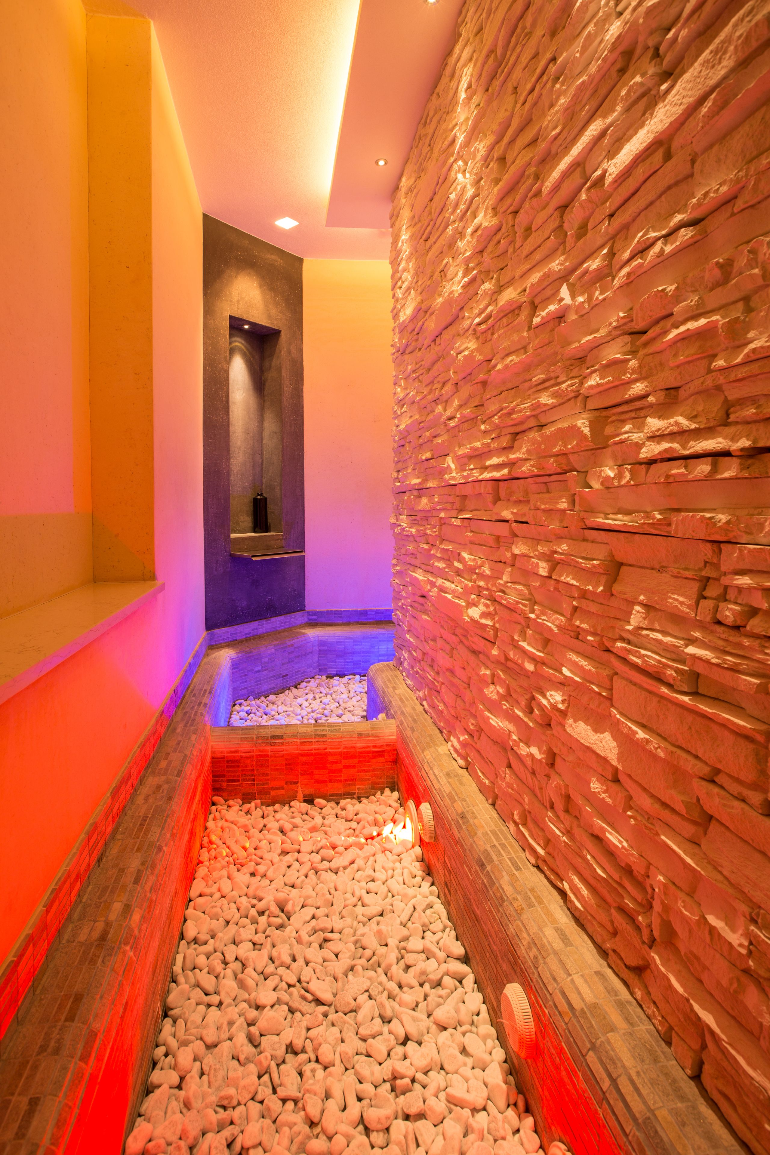 A narrow, dimly lit hallway with a pebble-lined floor, a stone wall on one side, and a bench in the distance.