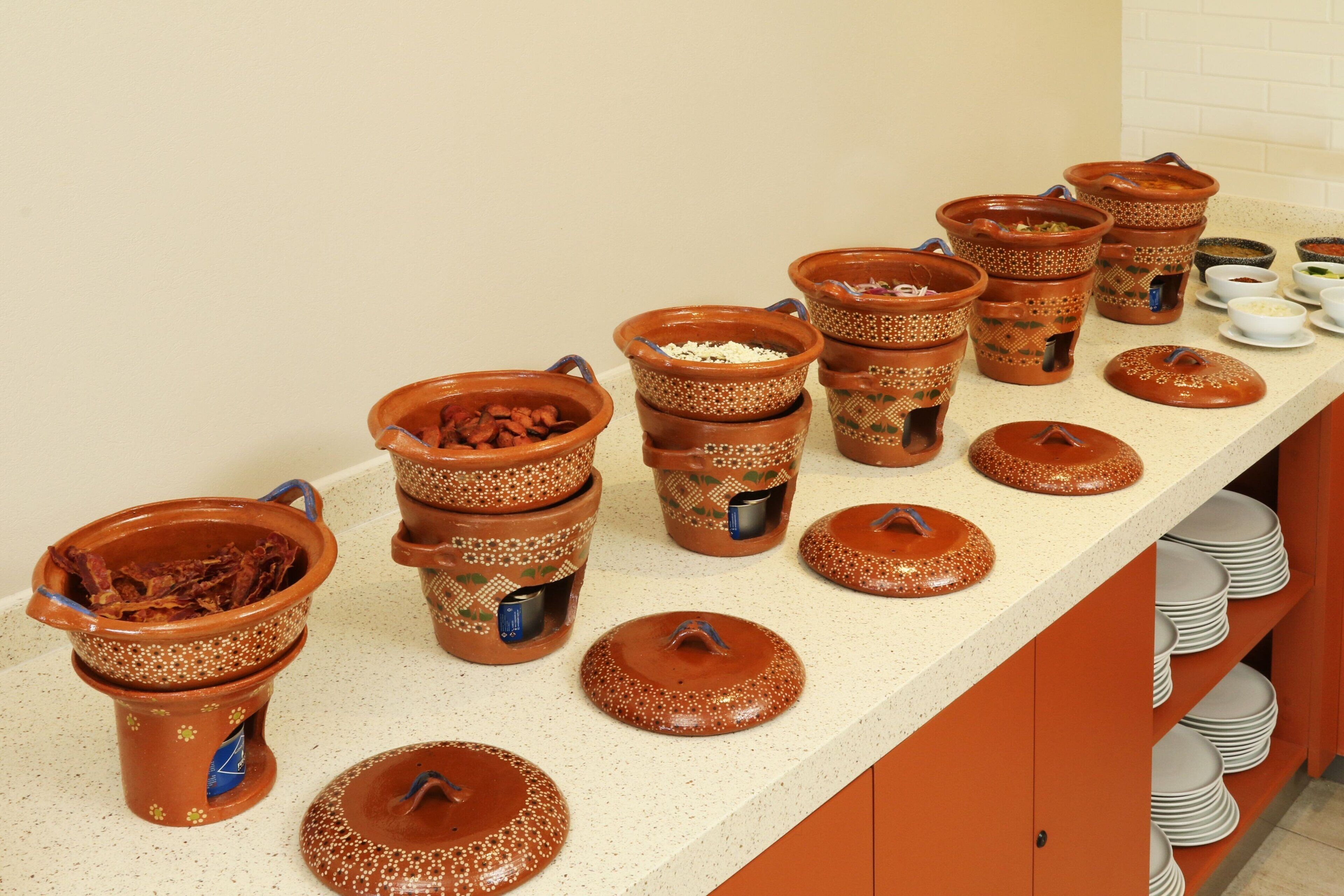 A kitchen counter with various ceramic pots and lids, stacked plates, and a tiled backsplash.