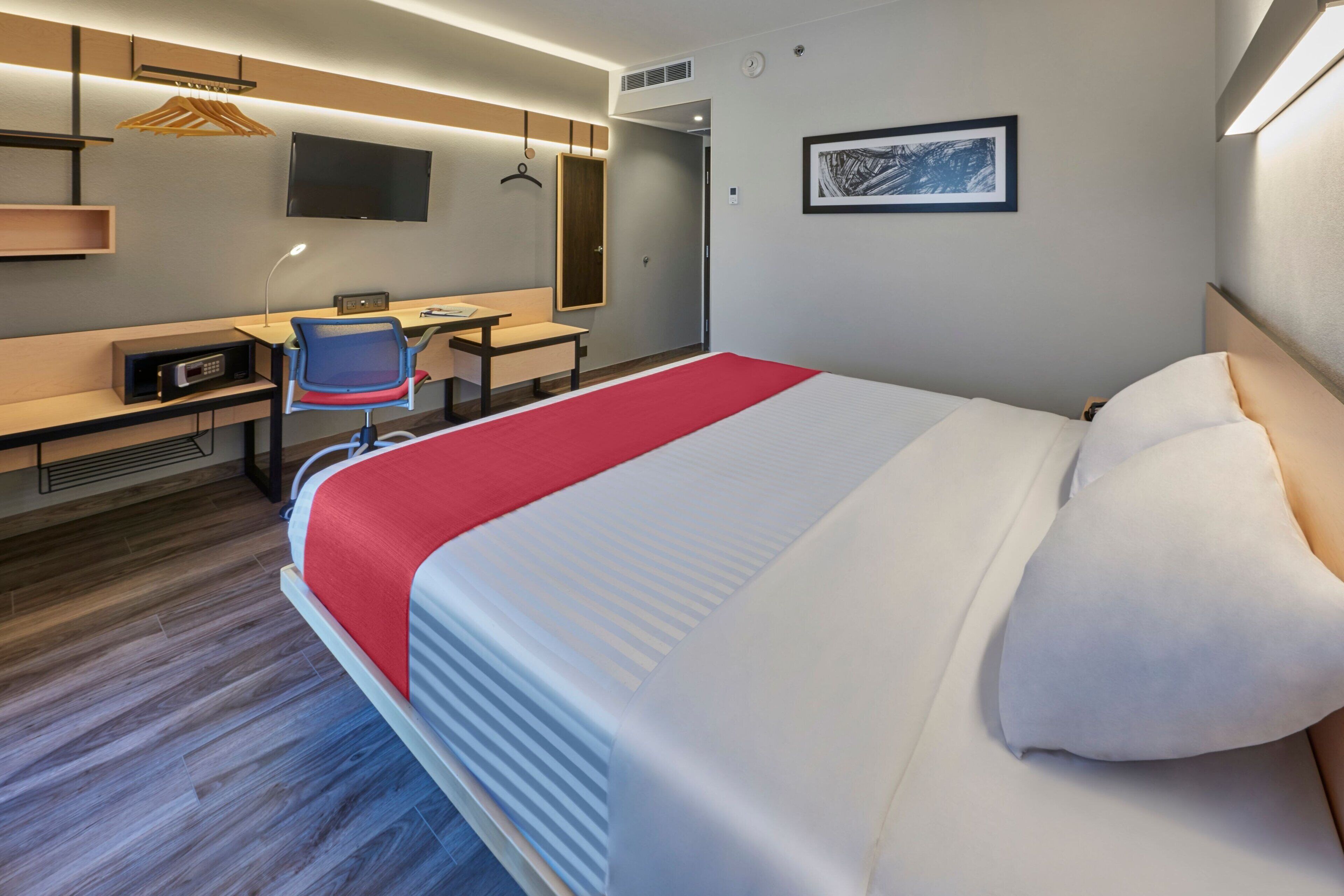 A modern hotel room with a large bed, a desk, a chair, a TV, and a red blanket.