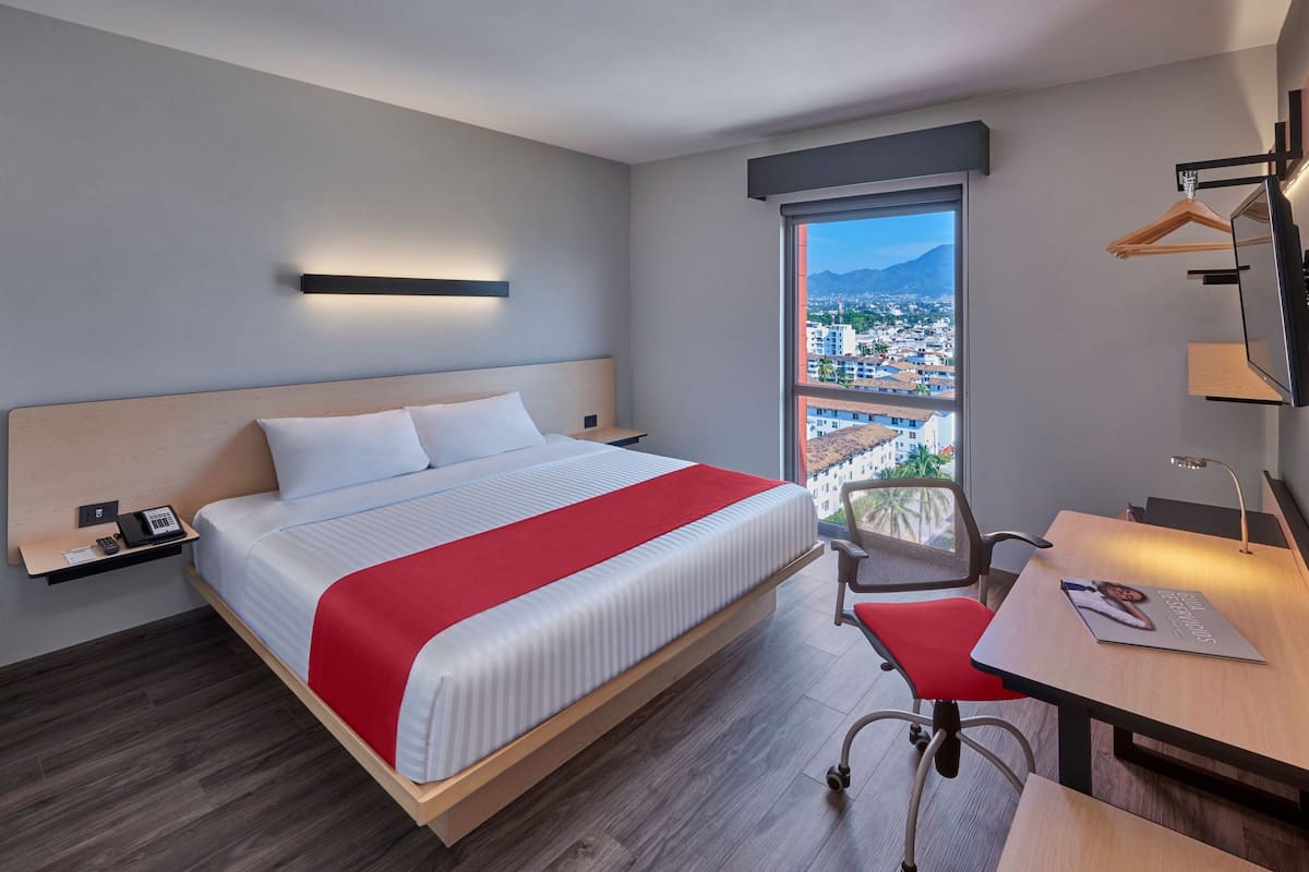 A modern hotel room with a large bed, a desk, a chair, a TV, and a window with a city view.
