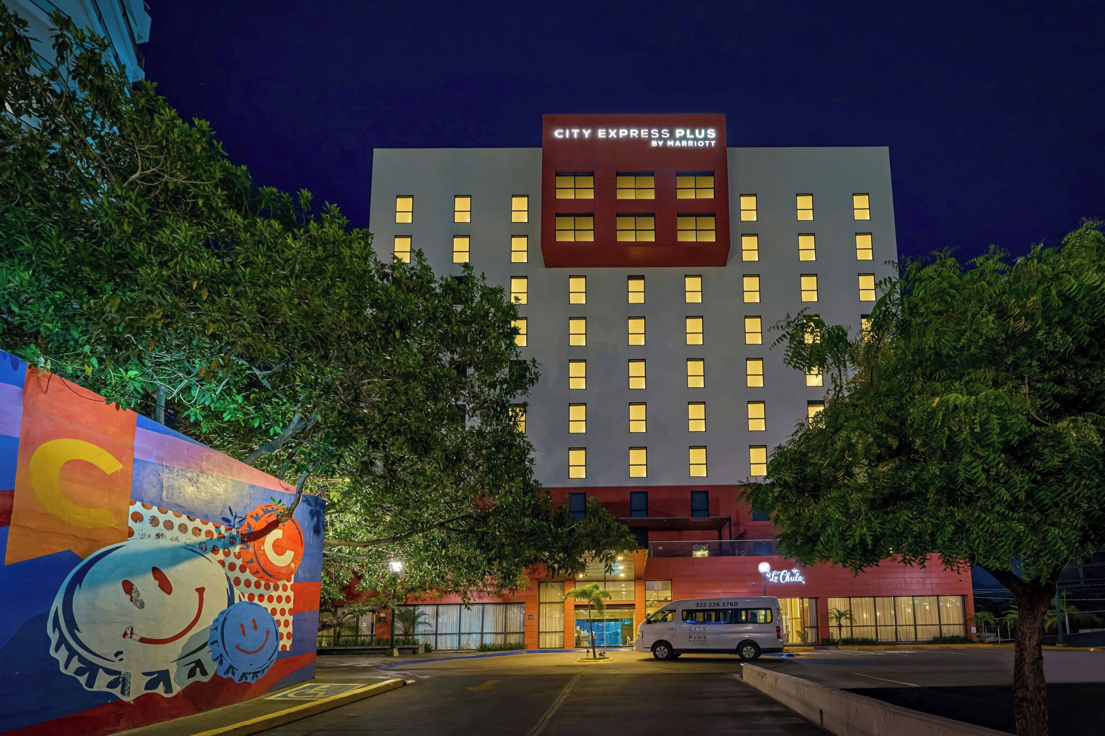 A hotel with a sign reading 'CITY EXPRESS PLUS BY MARRIOTT,' a parking area, and a mural on the left.