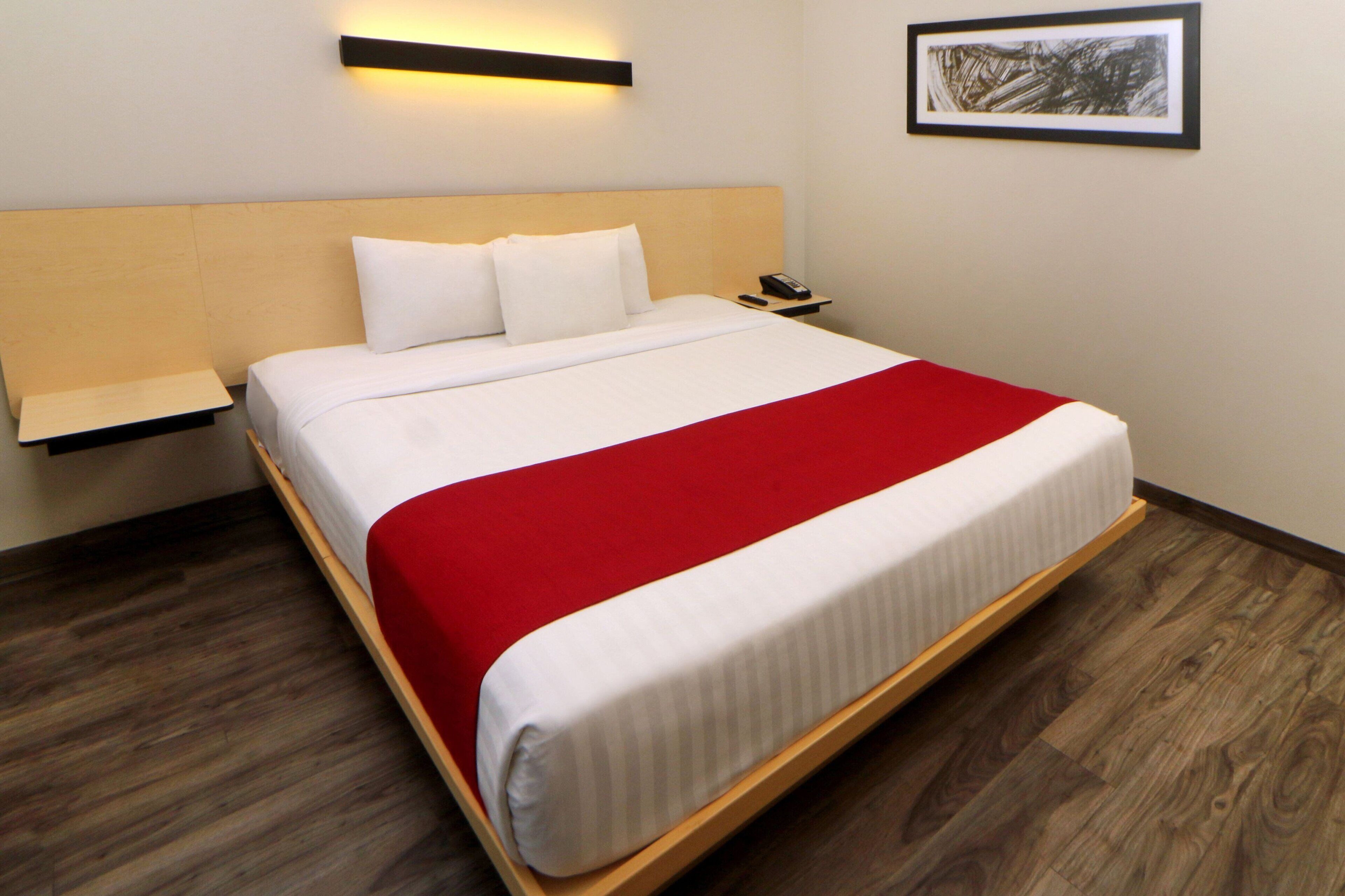 Ein ordentlich bezogenes Bett mit roter Decke, ein Nachttisch mit Telefon und ein gerahmtes Bild an der Wand.