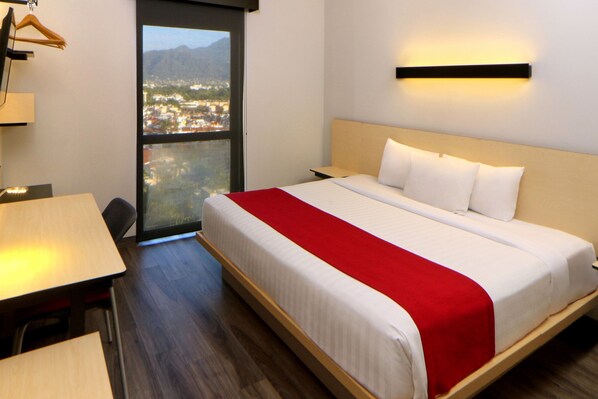 A hotel room with a bed, a desk, and a view of a cityscape through a large window. - City Express Plus by Marriott Puerto Vallarta (Puerto Vallarta)