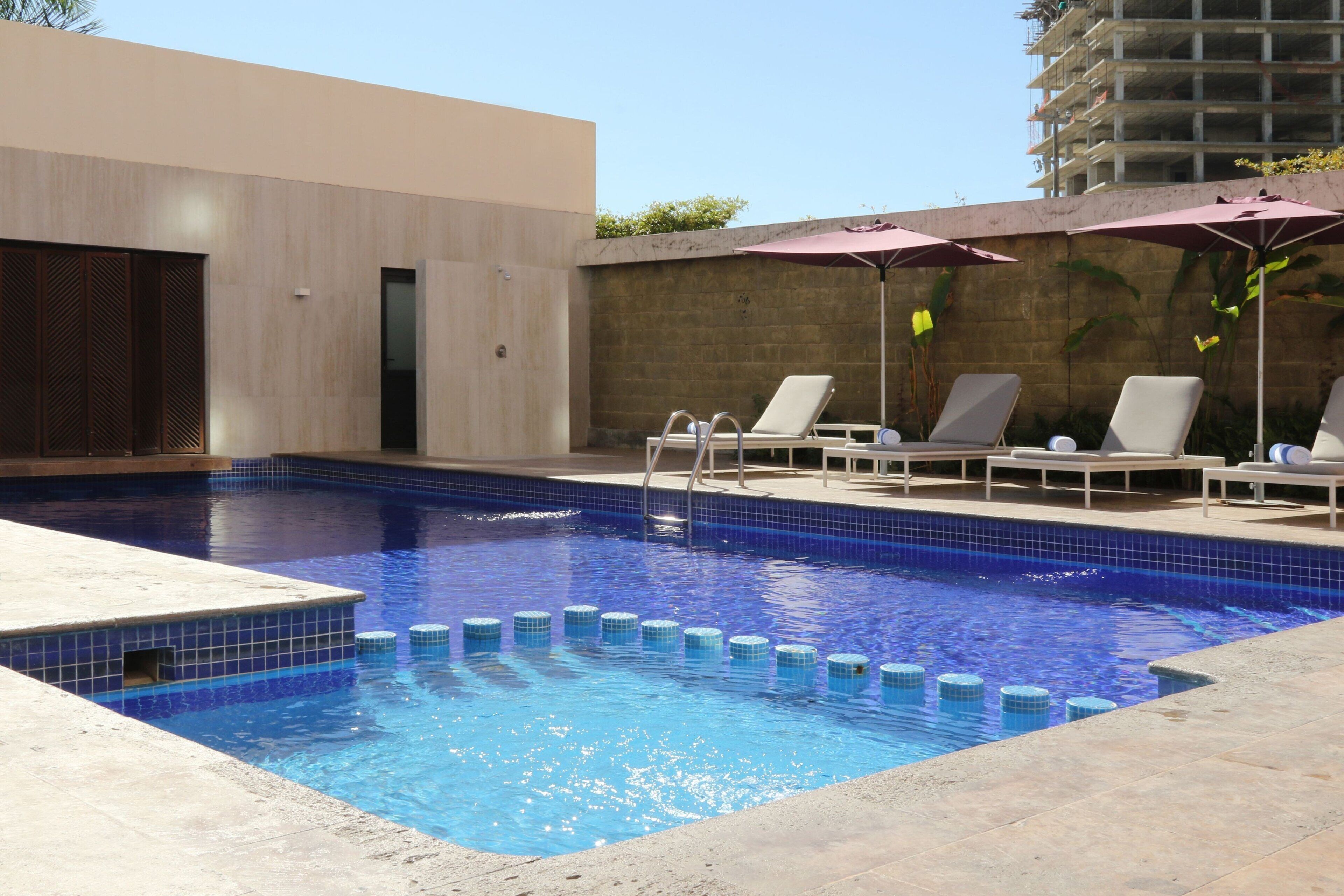 Une piscine avec des chaises longues et des parasols, un mur en bois et un bâtiment en arrière-plan.