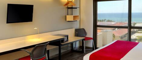 A hotel room with a bed, a desk, a chair, a TV mounted on the wall, and a view of the ocean through large windows.