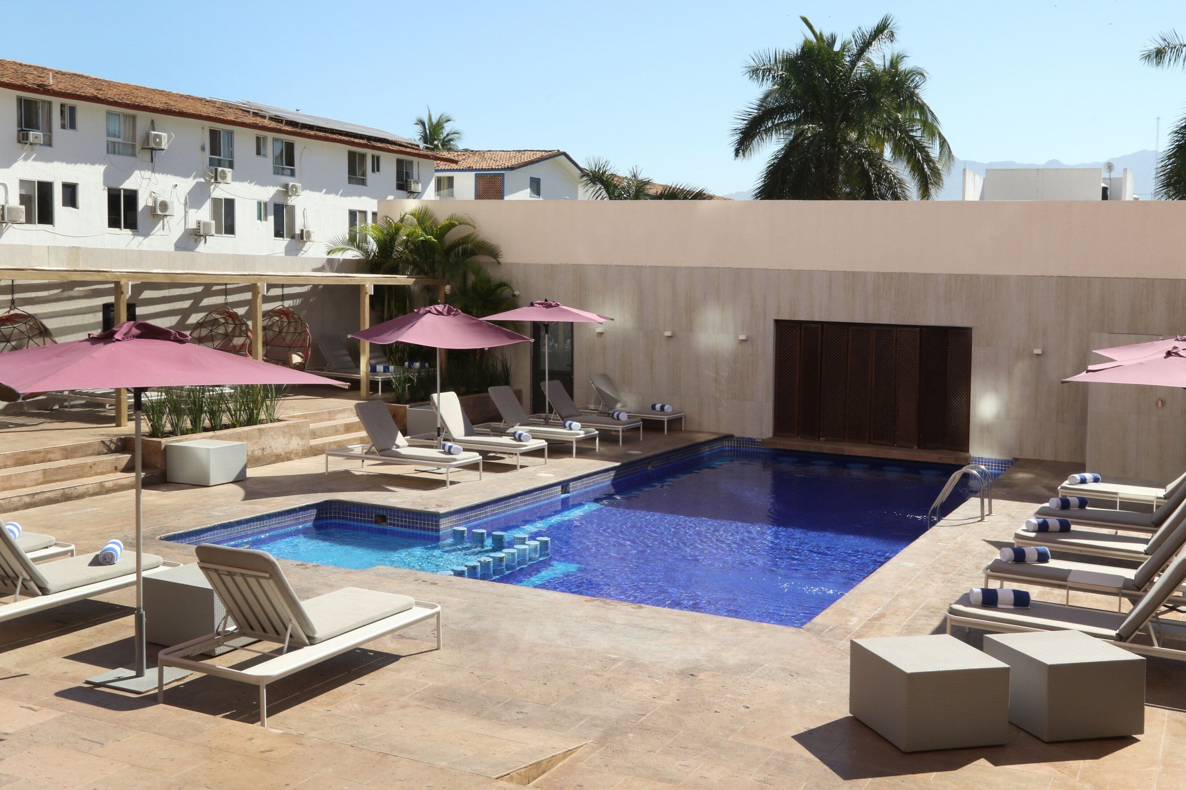Zona de piscina con sillones reclinables y sombrillas, piscina con baldosas y un edificio con múltiples ventanas y unidades de aire acondicionado.