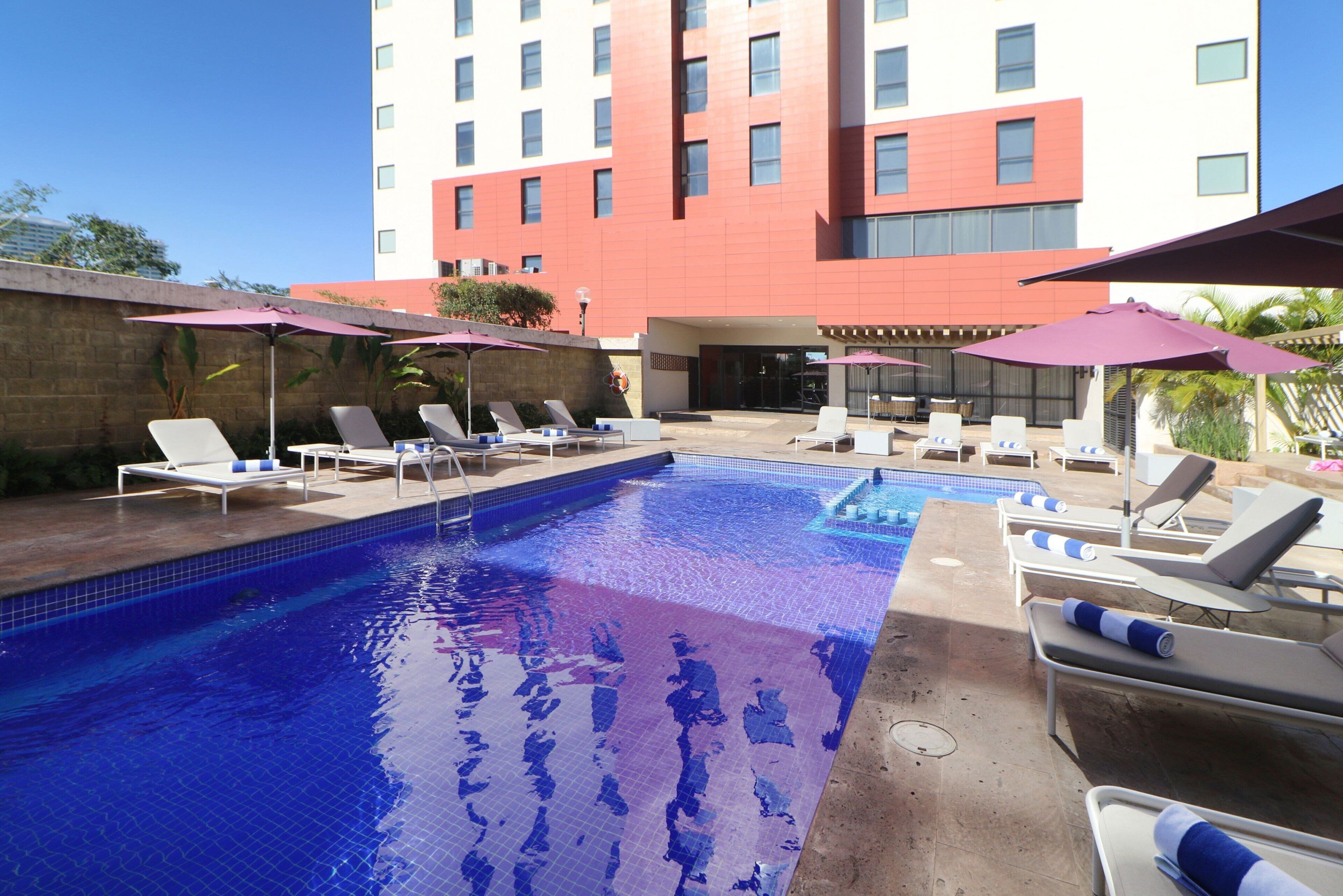 A swimming pool with lounge chairs and umbrellas, surrounded by a brick wall and a building with multiple windows.