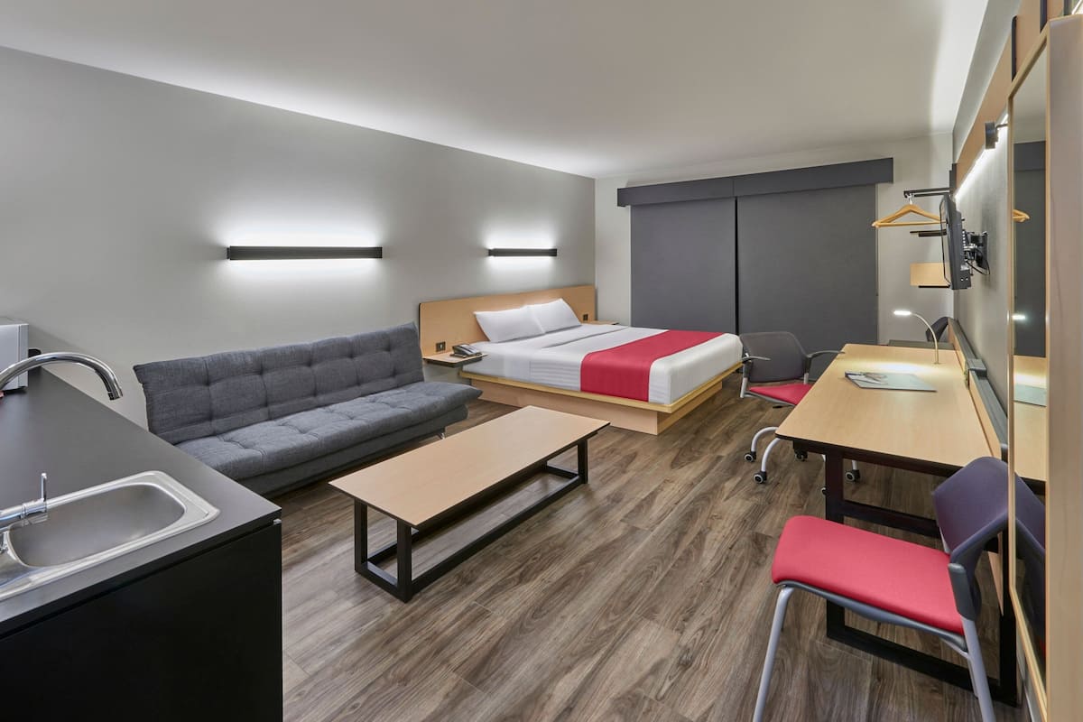 A modern hotel room with a sofa, bed, desk, and kitchenette.