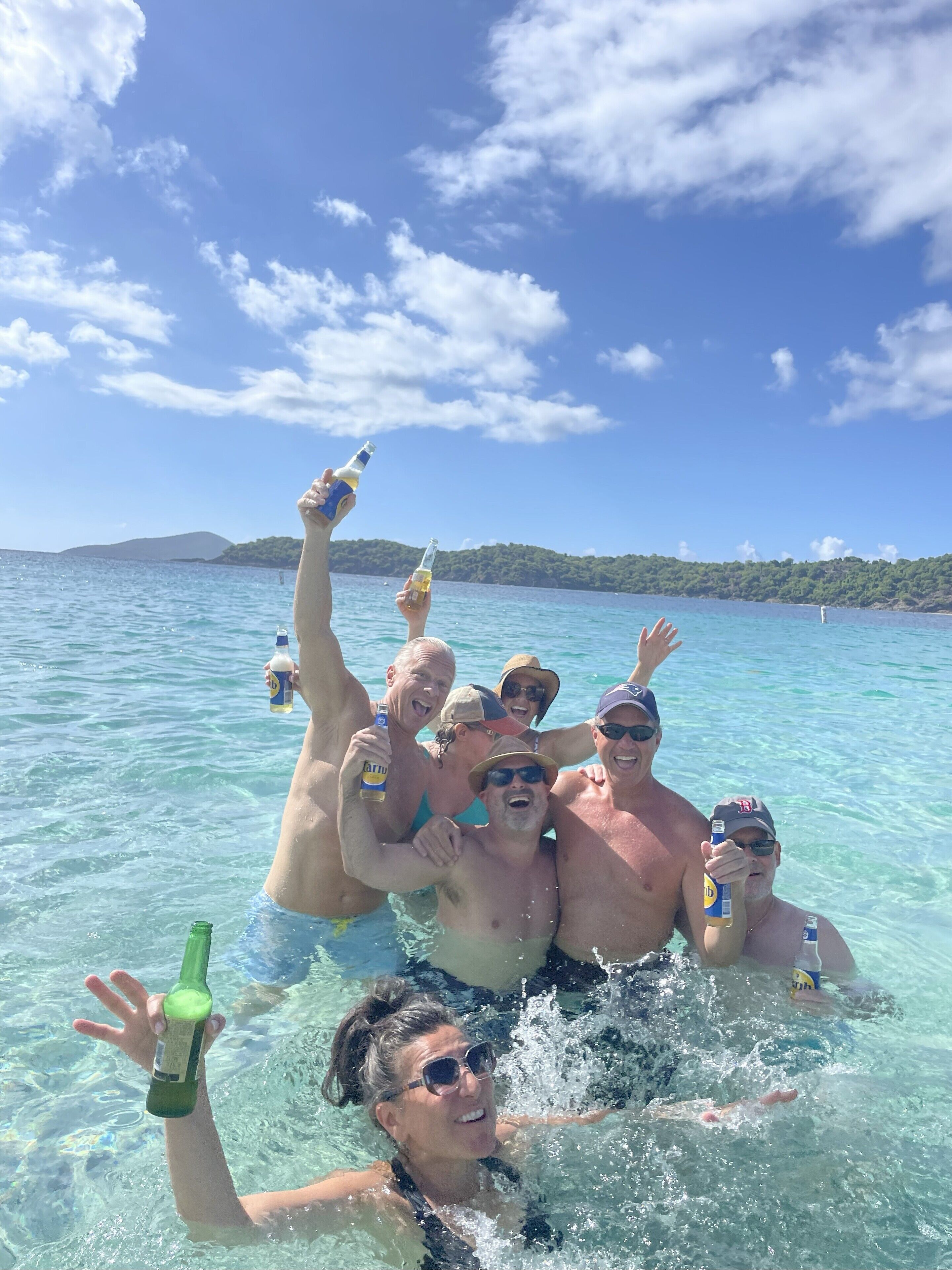 Un groupe de personnes se trouve dans une étendue d’eau d’un bleu limpide, tenant des bouteilles, avec un paysage montagneux en arrière-plan.