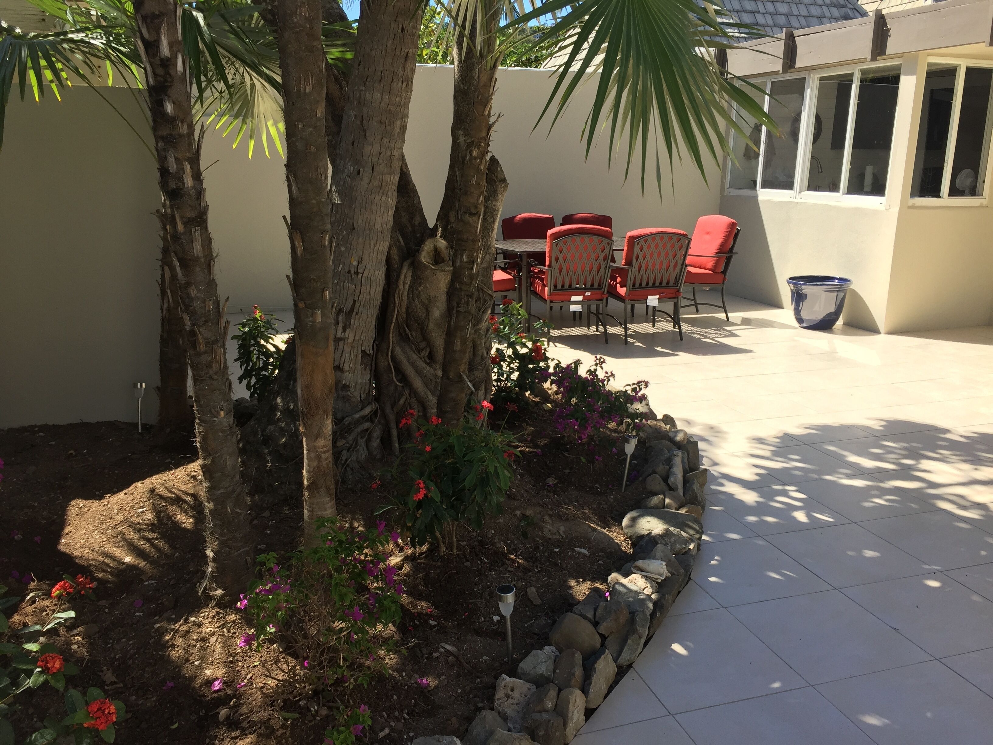 Eine Terrassenfläche mit Tisch und Stühlen, umgeben von einem Garten mit Palmen und blühenden Pflanzen.