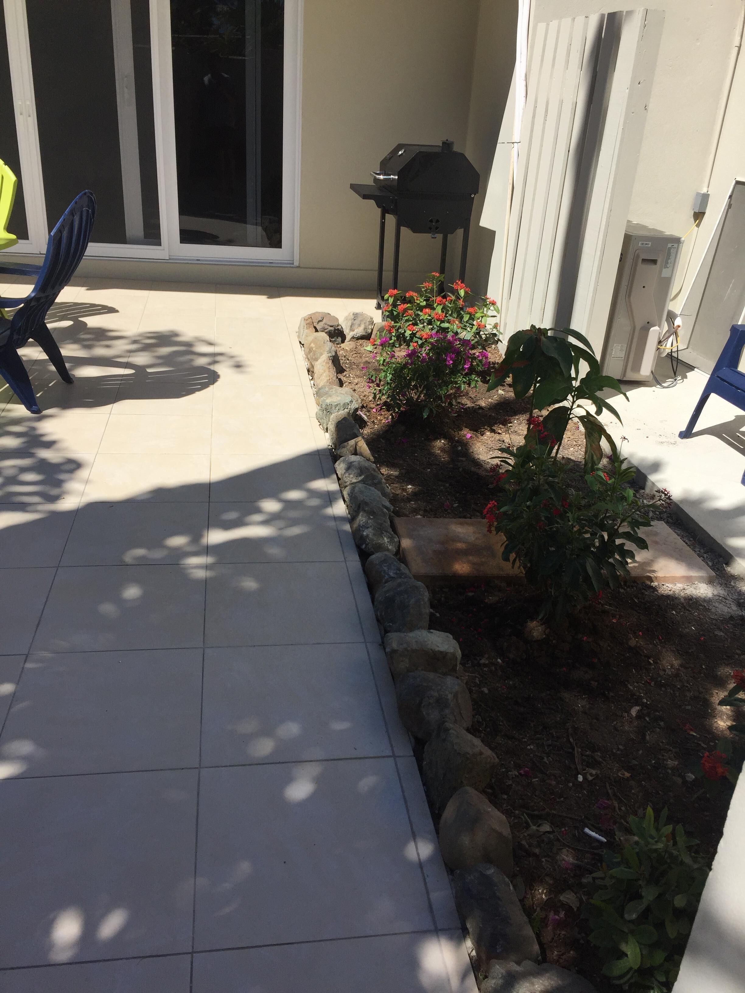 A patio with a tiled floor, a small garden with colorful flowers, a barbecue grill, and a blue chair.
