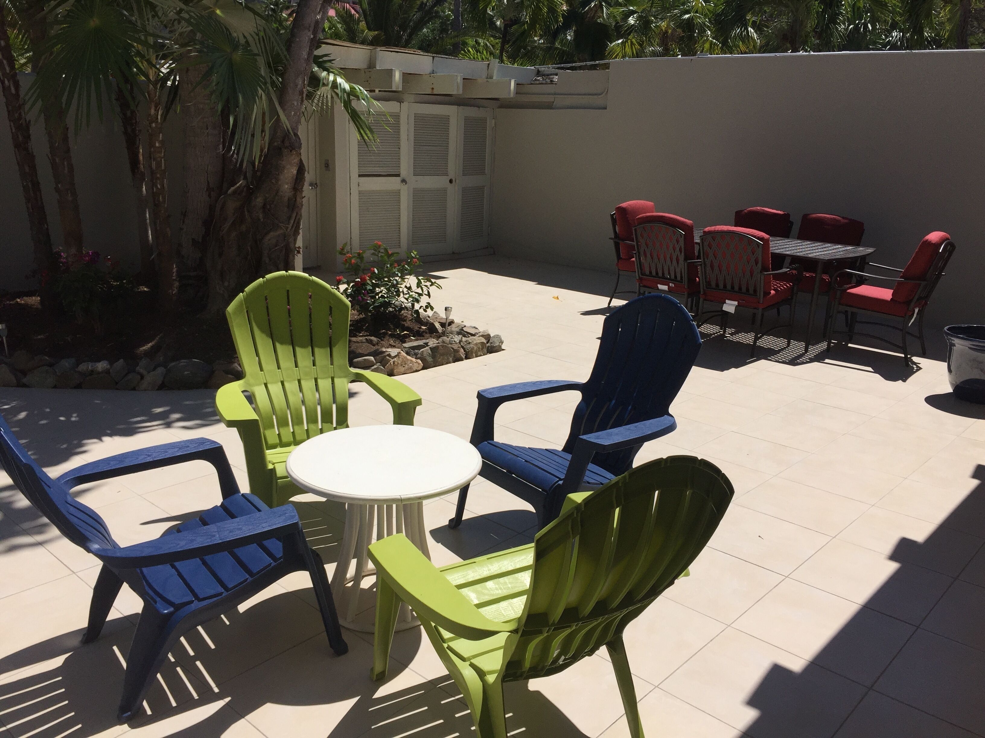 Une terrasse extérieure avec des chaises vertes et bleues, une table blanche et, au fond, des chaises rouges.