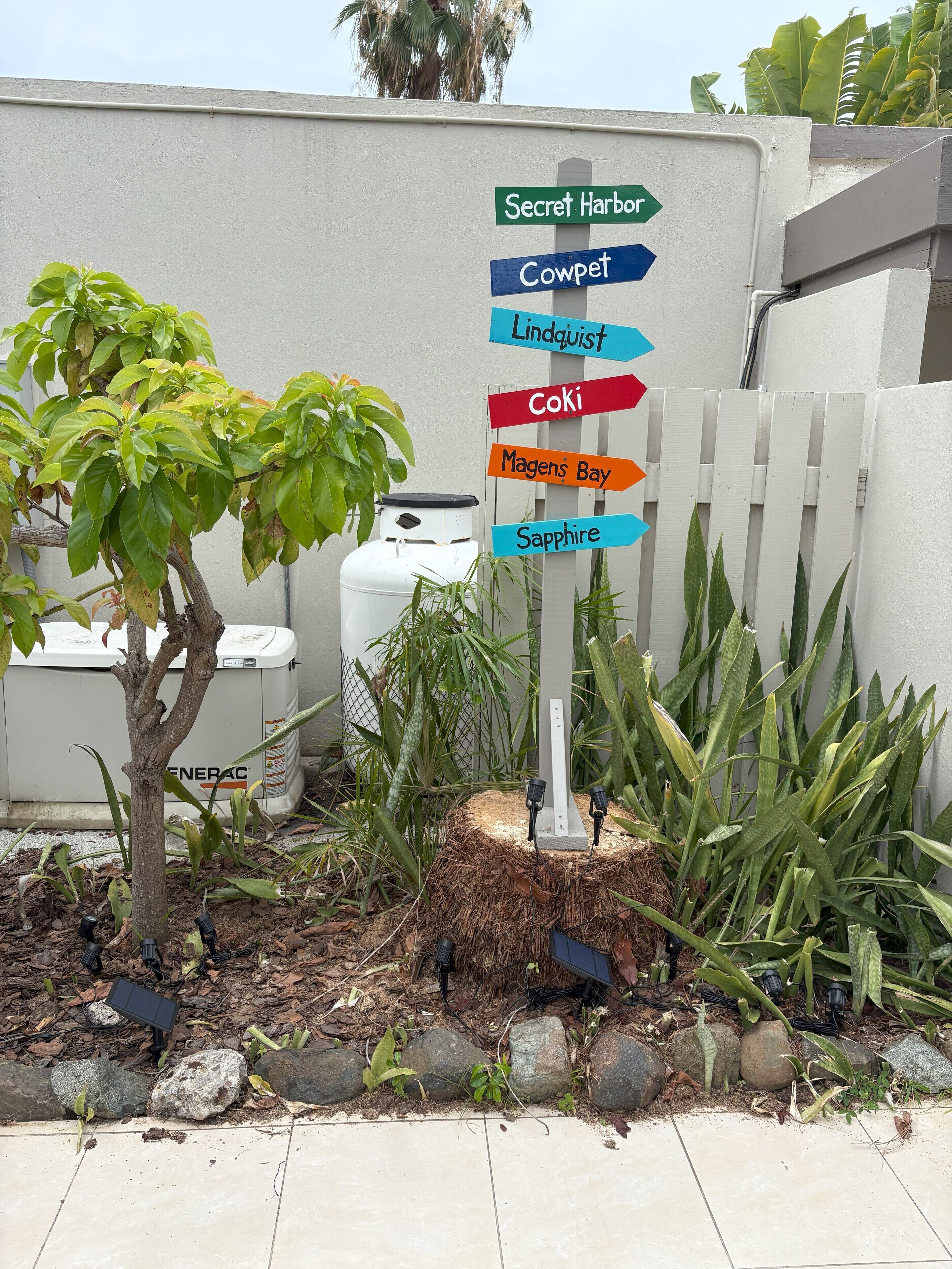 A directional signpost with multiple destinations labeled, surrounded by a garden with various plants and a white fence.