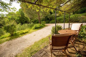A covered outdoor seating area with a table and chairs, surrounded by greenery and a gravel path.