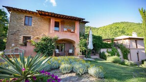 Una casa de piedra con balcón, un jardín con flores amarillas y una colina al fondo.