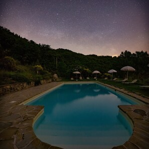 A swimming pool with a starry night sky above, surrounded by greenery and outdoor seating.