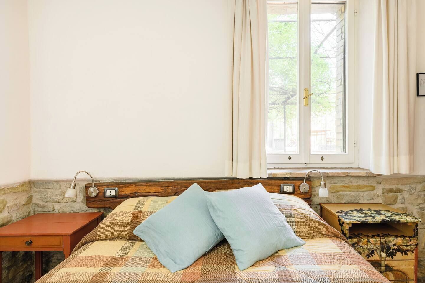 A bedroom with a bed, a nightstand, a lamp, a window with curtains, and a stone wall.