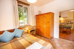 A bedroom with a bed, a wooden wardrobe, a window with curtains, and a view of a kitchen.