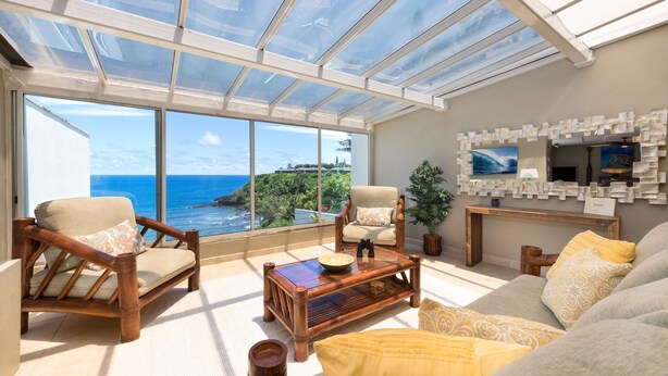 A spacious living area with a large glass window offering a view of the ocean and greenery. There are two wooden chairs, a sofa, and a coffee table with decorative items. - Premier Location, Huge Puu Poa Condo Ocean Views, Walk to 2 Beaches, Pool/Tennis (Princeville)