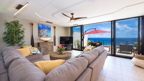 A modern living room with a large sofa, a television, and a view of the ocean through large windows. - Premier Location, Huge Puu Poa Condo Ocean Views, Walk to 2 Beaches, Pool/Tennis (Princeville)
