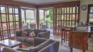 A spacious living area with a sofa and chairs, a kitchen counter, and large windows offering a view of the outdoors. - POCO POCO Villa - Caribbean Golf, Diving & Beach Holiday Home (Blue Bay Golf & Beach Resort, Willemstad, Curacao)