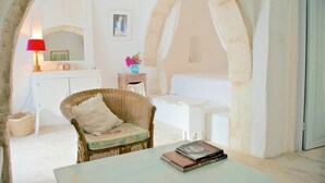 Habitació amb una cadira de vimet, una taula petita amb llibres, un sofà blanc i un mirall decoratiu.