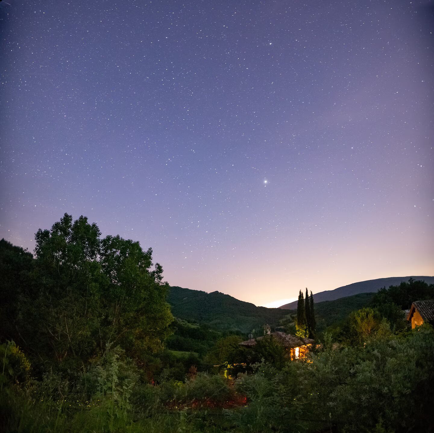 Cielo estrellado sobre un paisaje rural, con una pequeña casa iluminada al frente.