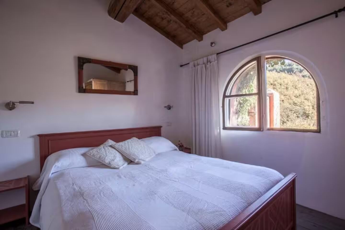 A bedroom with a wooden bed, a large window with a view of greenery, a mirror on the wall, and a wooden ceiling.