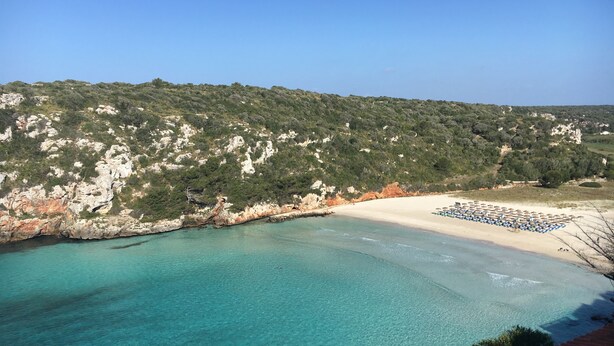 Una spiaggia costiera con acque turchesi cristalline, una riva sabbiosa e un pendio roccioso sullo sfondo.