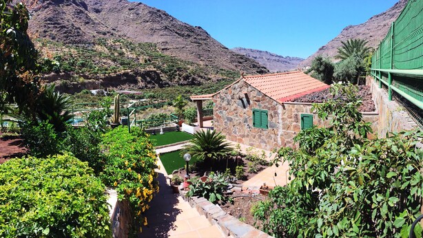 A stone building with a red-tiled roof, surrounded by lush greenery and a mountainous backdrop. - TINO HOUSE - FOR 5 PEOPLE- SWIMMING POOL (Mogán)