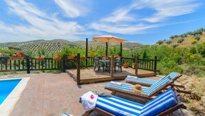 Un espace aménagé au bord de la piscine, avec des chaises longues et une table placée sous une pergola, le tout entouré d’une terrasse en bois et donnant sur la piscine.