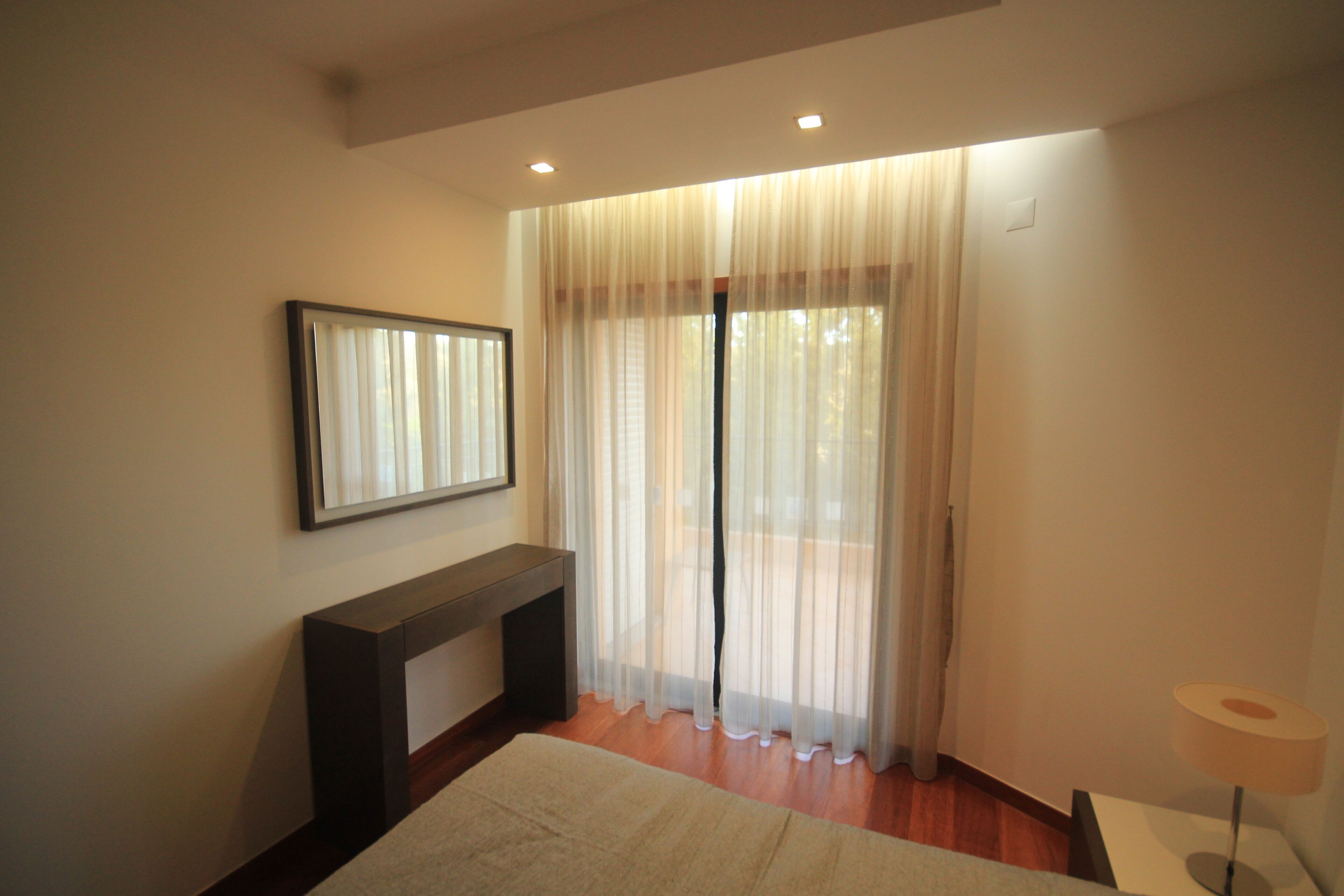 Un dormitorio con cama, mesita de noche, lámpara y ventana con cortinas translúcidas.