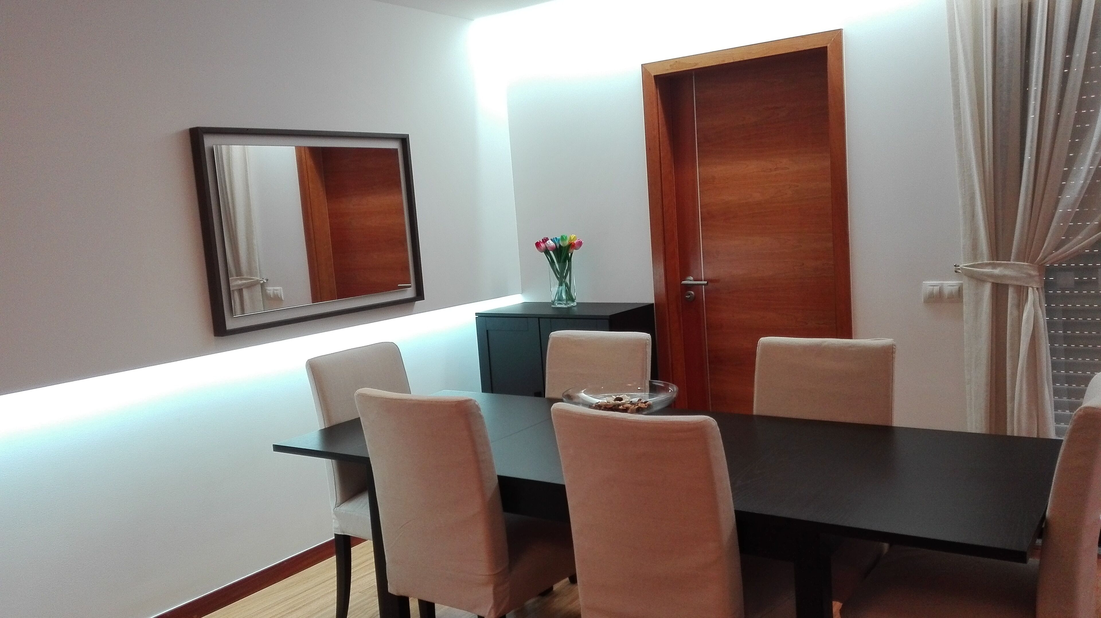 A dining area with a dark table, light-colored chairs, a vase with flowers, and a mirror on the wall.