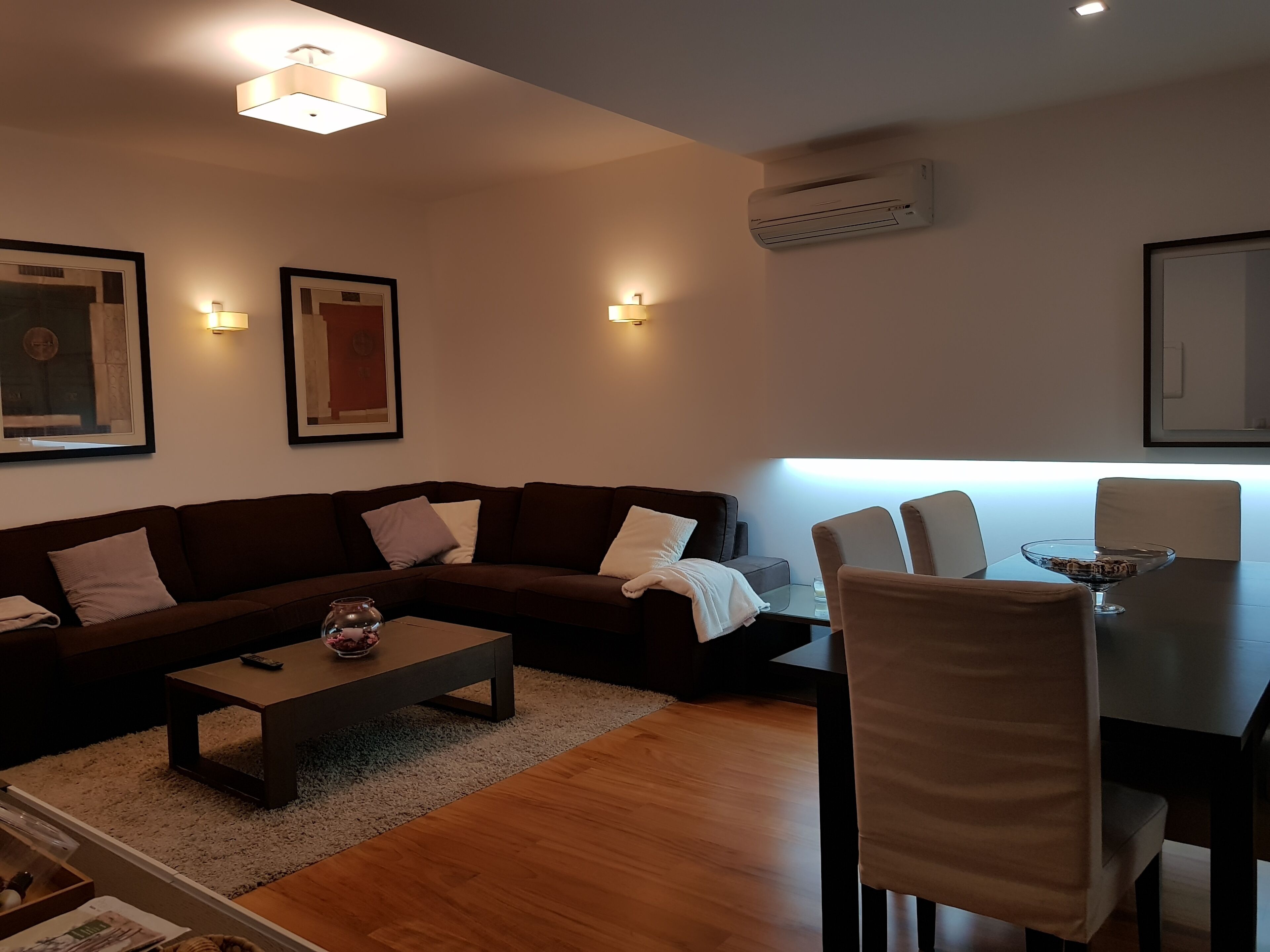 A modern living room with a brown sofa, a coffee table, and a dining table with chairs. There are wall-mounted lights and an air conditioning unit.