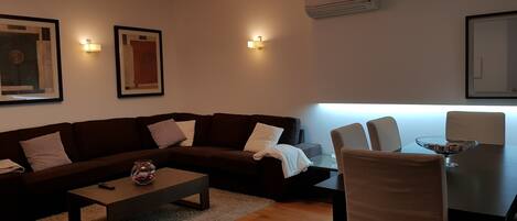 A modern living room with a brown sofa, a coffee table, and a dining table with chairs. There are wall-mounted lights and an air conditioning unit.