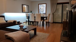 Un salon moderne avec un canapé, une table basse et un meuble pour la télévision. On y trouve également un coin repas avec des chaises et une table, et une cuisine visible à travers une porte ouverte.