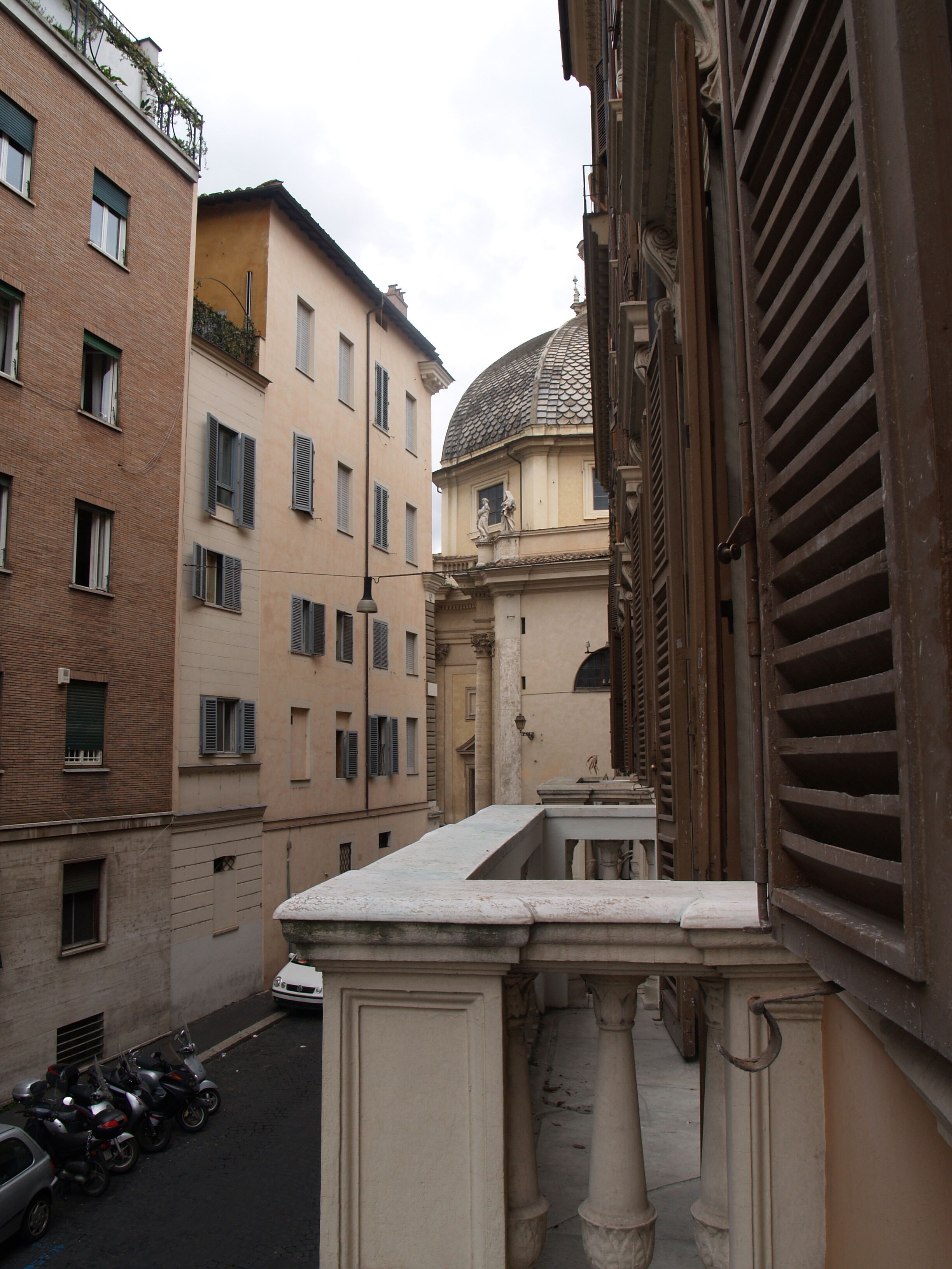 A narrow street with buildings on either side, a row of parked scooters, and a small balcony with a stone balustrade.
