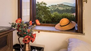 Ein Zimmer mit Fensterblick auf eine malerische Landschaft, einem Holzkasten, einer Blumenvase und einem Strohhut auf der Fensterbank.