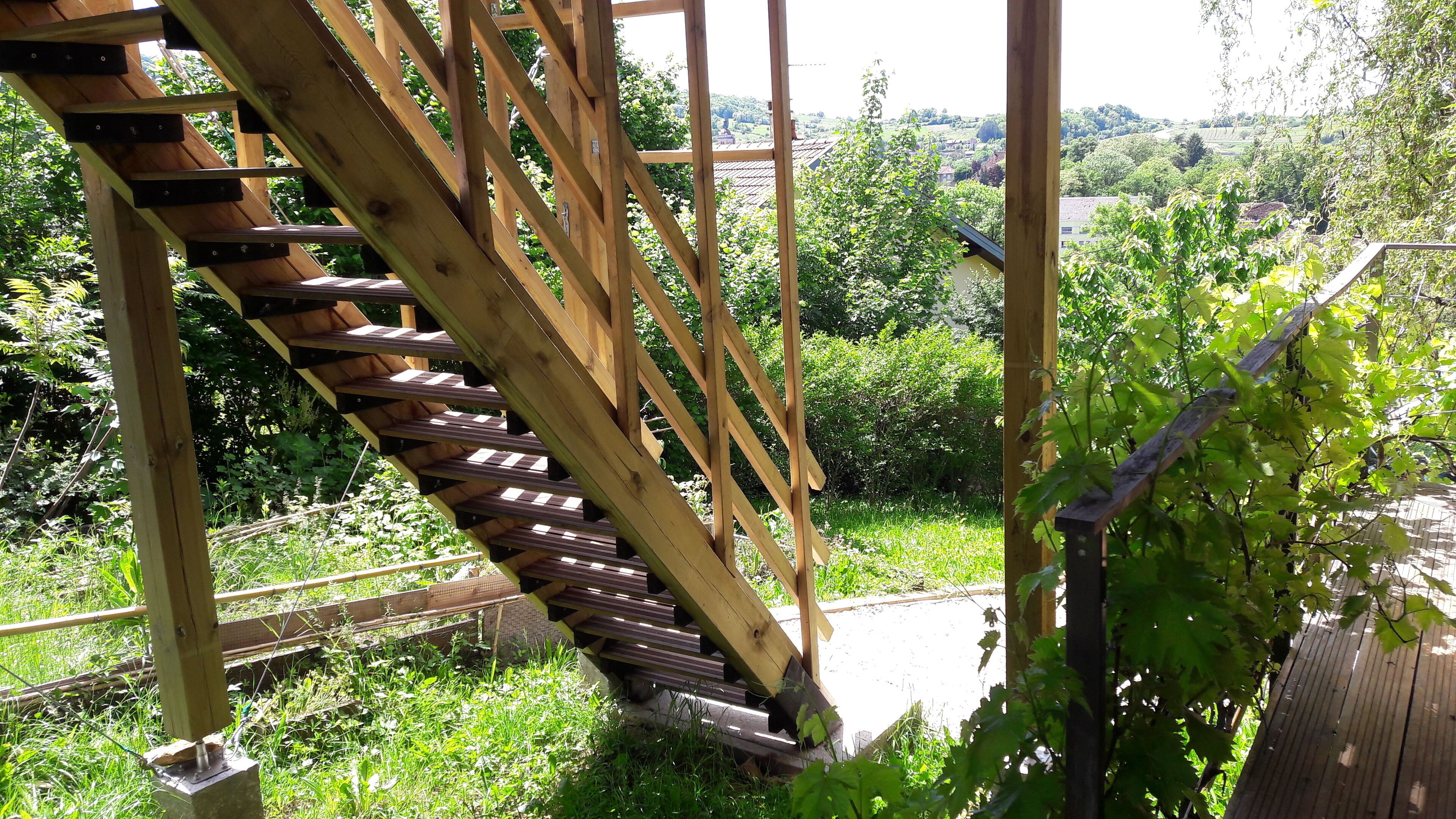 A wooden spiral staircase leading to a garden area with lush greenery and a view of a rural landscape.
