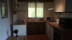 A compact kitchen with a sink, microwave, stove, and a small window with curtains. - Gite au figuier - studio Duplex 2/4 people (Arbois)
