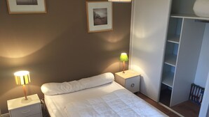 Ein ordentlich bezogenes Bett mit weißer Bettwäsche, zwei Nachttischlampen, eine kleine Schublade am Nachttisch und ein an der Wand befestigtes Regal mit einem gerahmten Bild.