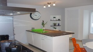 Eine moderne Küche mit Holzarbeitsplatte, Spüle und einer Uhr an der Wand. Es gibt Schränke, ein Regal mit Pflanzen und ein Fenster mit Jalousien.