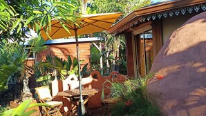 Een terras buiten met een houten tafel en stoelen, een gele parasol en weelderige tropische planten.