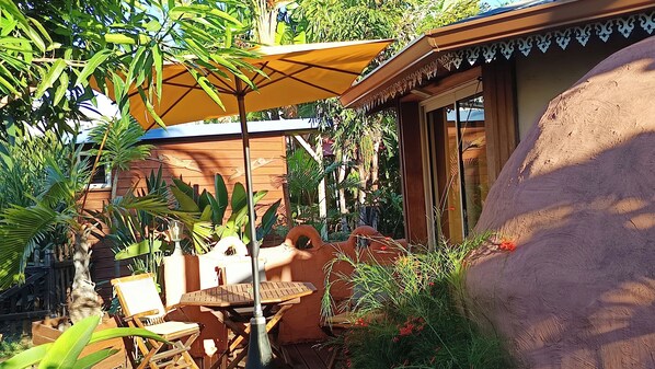 An outdoor patio with a wooden table and chairs, a yellow umbrella, and lush tropical plants.