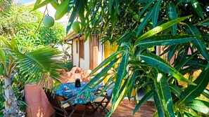 A wooden deck with a table and chairs, surrounded by lush greenery and a small building. - Ti Caze Dodo, small Creole hut (Etang-Salé les Hauts)