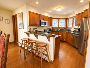 A modern kitchen with wooden cabinets, a stainless steel refrigerator, and a bar area with stools. - C7 Beautifully appointed slopesideCrawford Ridge end unit townhome with fireplaces, yard (Bretton Woods)