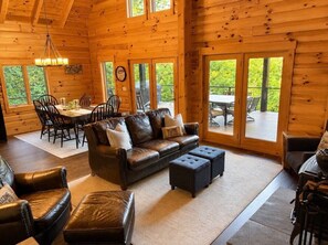 A log cabin interior with a leather sofa, ottomans, dining table, chairs, and large windows.