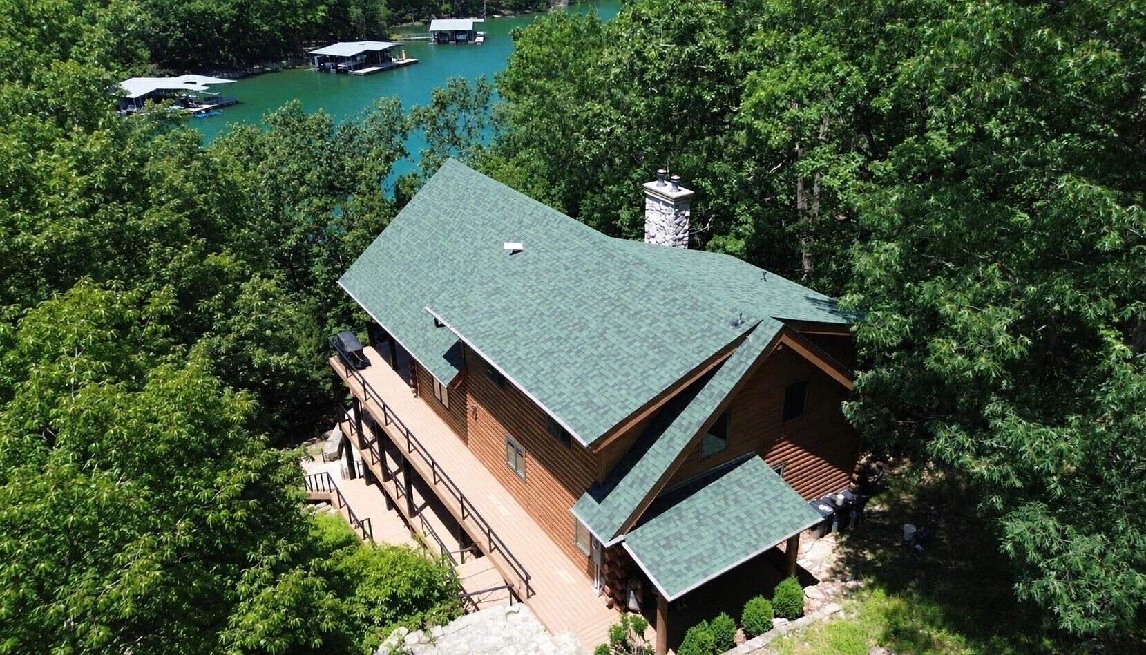 A cabin with a green roof, surrounded by dense trees, and a dock extending into a lake.