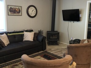 Un salon avec un canapé noir, un fauteuil marron, un téléviseur noir fixé au mur et un poêle à bois noir.